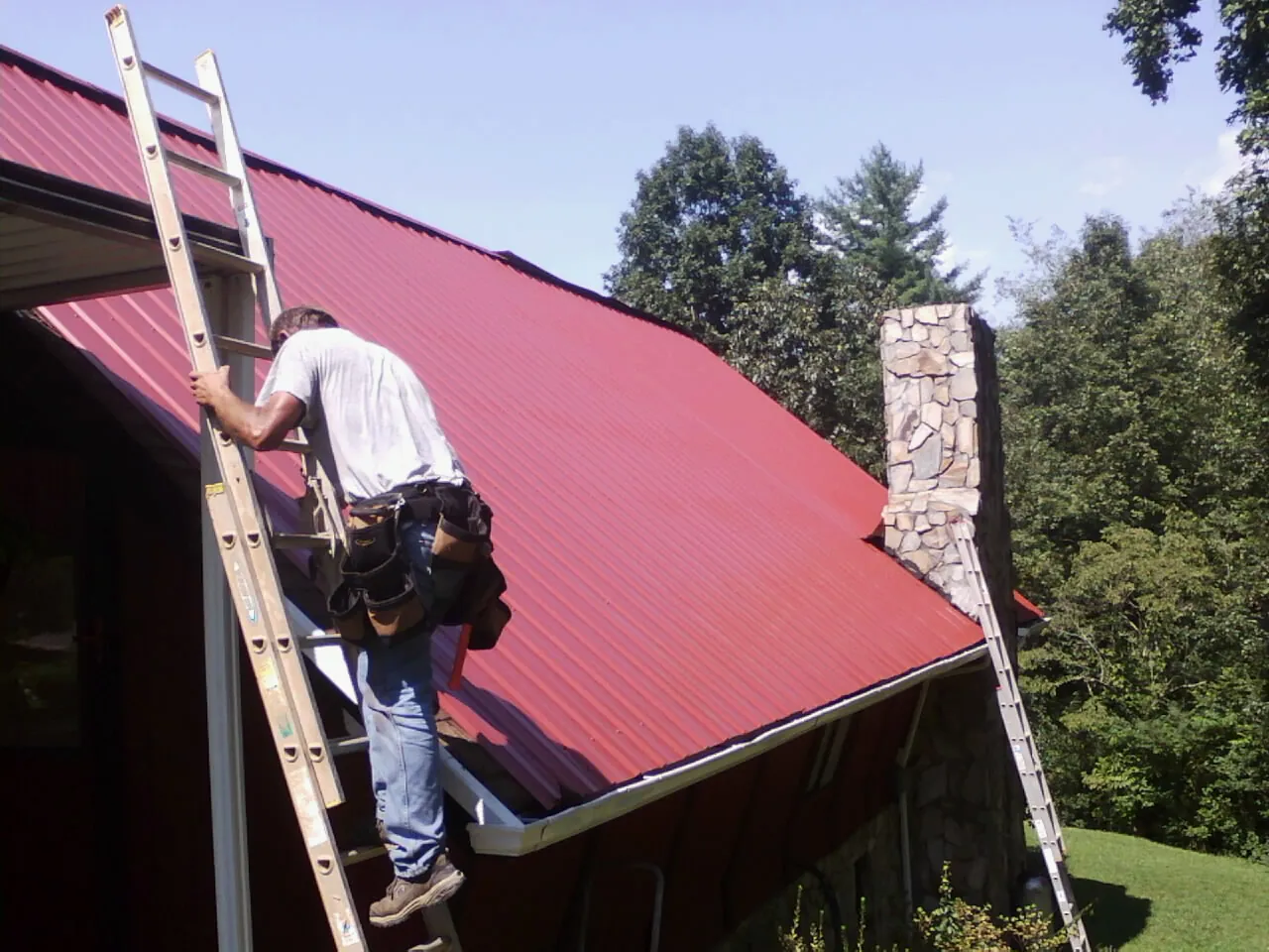 Roofer on a ladder beside a red metal roof and stone chimney — Soffit & Fascia Repair by Roofing Brothers in Platte Center